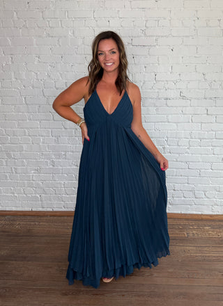 A woman modeling a deep V-neck, floor-length pleated maxi dress in dark teal blue, standing against a white painted brick wall on a wooden floor.