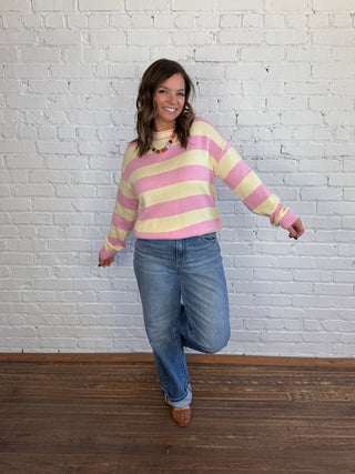 A woman wearing a pink and yellow striped knit sweater, blue jeans, and brown shoes, posing against a white brick wall and dark wood floor.