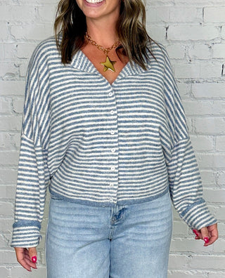 A woman modeling a long-sleeve striped button-up shirt and light-wash jeans, standing against a white brick wall on a wooden floor.