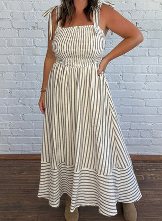 A woman modeling a sleeveless, square-neck maxi dress with vertical stripes in cream and taupe, standing against a white brick wall on a dark wood floor.