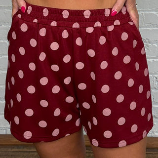 A woman modeling a matching burgundy and pink polka dot two-piece set standing against a white brick wall and wooden floor.
