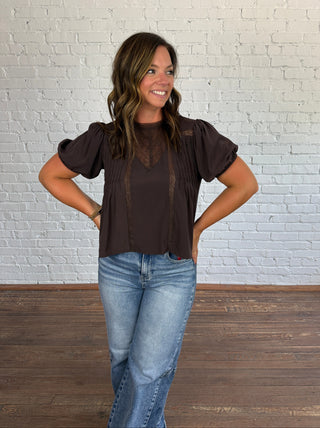 A woman modeling a dark brown short-sleeve blouse with sheer lace panels on the chest and sleeves, paired with medium wash blue jeans, standing against a white brick wall.