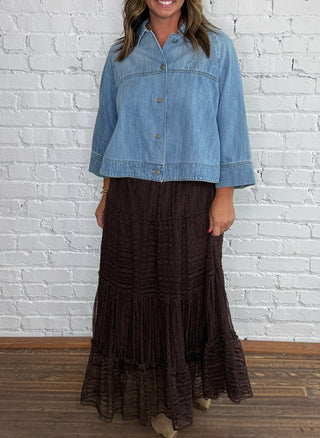 A woman stands in front of a white brick wall wearing a cropped denim top and a dark brown tiered lace midi skirt, paired with taupe ankle boots.
