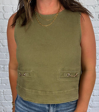 A woman stands in a room with a white brick wall and dark wood floor, wearing a sleeveless olive green top with gold equestrian horse bit faux pocket detail and wide-leg blue jeans.