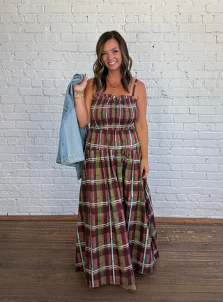 A woman stands in front of a white brick wall wearing a long plaid maxi dress with thin straps, holding a light blue denim jacket over her shoulder.