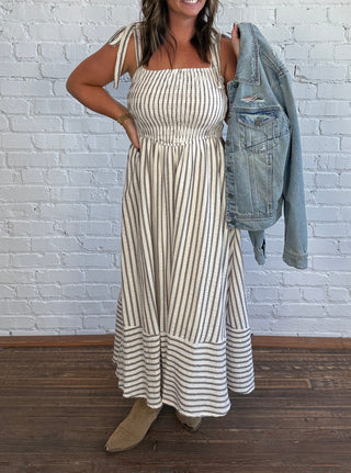 A woman stands in front of a white brick wall wearing a cream and taupe vertically striped maxi dress with a smocked bodice and ruffled hem, holding a light blue denim jacket over her shoulder.