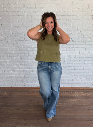 A woman stands in a room with a white brick wall and dark wood floor, wearing a sleeveless olive green top with gold equestrian horse bit faux pocket detail and wide-leg blue jeans.