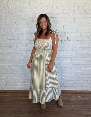A woman modeling a sleeveless, smocked maxi dress with vertical cream and green stripes, standing against a white brick wall on a wooden floor.
