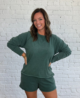 A woman modeling a matching dark green long-sleeve top and shorts set, standing against a white brick wall on a wooden floor.