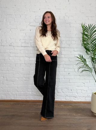 Woman wearing a cream sweater and black pants standing in front of a white brick wall with a plant to the right.