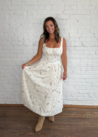 A woman modeling a spaghetti strap cream maxi dress with a delicate floral pattern, standing against a white brick wall on a dark wood floor.