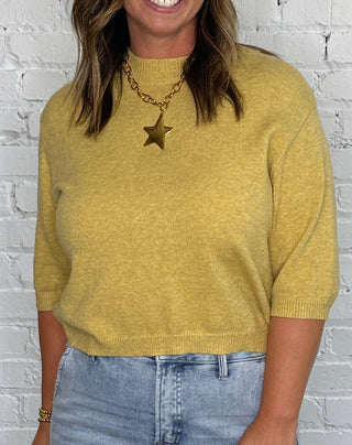 A woman stands in front of a white brick wall wearing a mustard yellow knit sweater, light blue jeans, and tan pointed-toe heels.