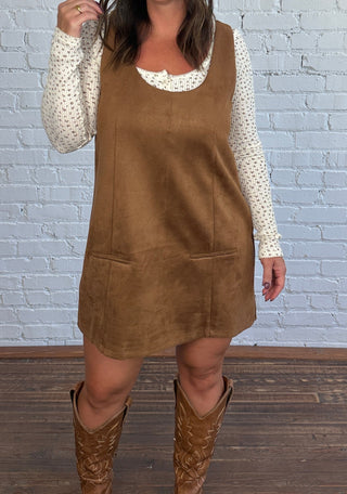 A woman modeling a brown dress over a cream, floral, long-sleeve top, paired with brown cowboy boots, standing against a white brick wall.