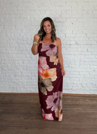 A woman wearing a strapless maxi dress with a large floral print in burgundy, pink, purple and beige, standing against a white brick wall and wooden floor.