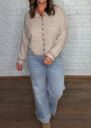 A woman stands in front of a white brick wall wearing a beige and brown striped button-up shirt and light blue jeans.