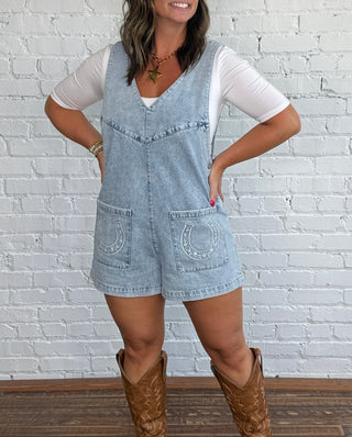 A woman stands in front of a white brick wall wearing a short, light blue denim overall romper with a horseshoe embroidered on each pocket, a white t-shirt underneath, and brown cowboy boots.