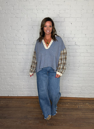 A woman modeling a mineral washed denim top with plaid sleeves and wide-leg blue jeans, standing against a white painted brick wall on a wooden floor.