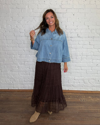 A woman stands in front of a white brick wall wearing a light blue cropped denim  button down top and a dark brown tiered midi skirt, paired with tan boots.