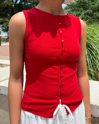 Woman wearing a red sleeveless top with buttons, standing outdoors with greenery in the background.