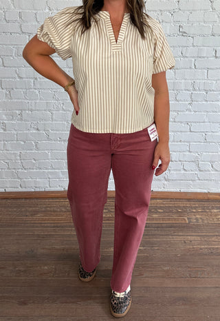 A woman modeling a cream and beige vertically striped short-sleeve blouse with puff sleeves, paired with dusty rose pants, standing in front of a white brick wall and wood floor.