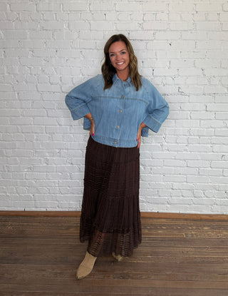 A woman posing in a light blue cropped denim jacket and a dark brown tiered lace midi skirt, standing on a wooden floor against a white brick wall.