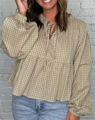A woman stands in front of a white brick wall wearing a beige and green gingham long-sleeve blouse with a tie-neck detail and light blue jeans.