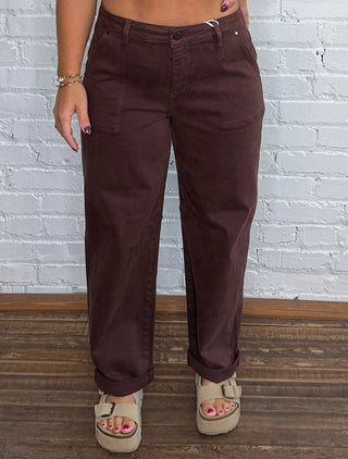 A woman modeling a pair of dark brown barrel jeans standing against a white brick wall on a wooden floor.
