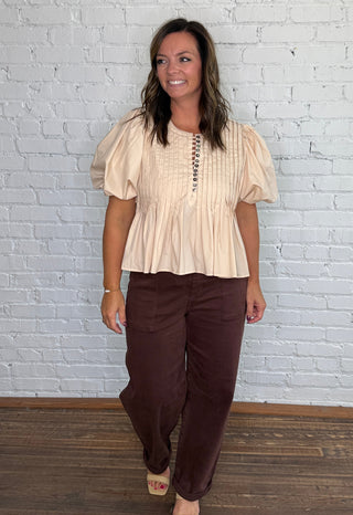 A woman posing in a cream puff-sleeve peplum top and dark brown barrel jeans against a white brick wall and wood floor.