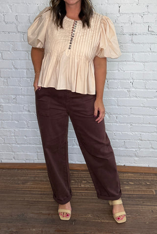 A woman posing in a cream puff-sleeve peplum top and dark brown barrel jeans against a white brick wall and wood floor.
