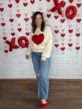 Woman wearing a cream sweater with a red heart and blue jeans standing in front of a heart-themed wall.
