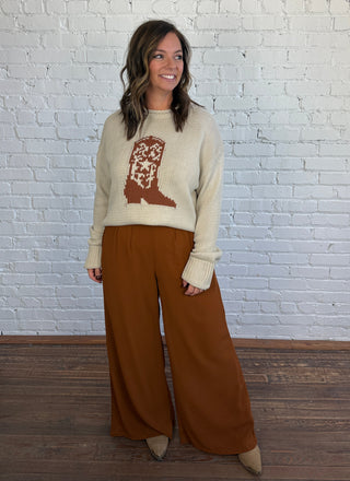 Woman wearing a beige sweater with a cowboy boot graphic woven into the sweater, paired with a pair of brown trousers and styled with top boots.