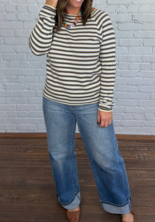 Woman wearing a cream sweater with baby blue and brown stripes, colorful necklace, denim, and brown flats.