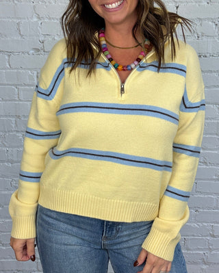 Women wearing a classic half-zip mock neck sweater in pale yellow with eye-catching blue stripes and delicate brown stripe paired with jeans and brown flats.