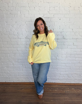 Woman wearing a butter yellow sweater with a blue and brown trout on the front paired with medium washed denim, cuffed jeans, and brown shoes.