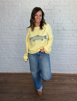 Woman wearing a butter yellow sweater with a blue and brown trout on the front paired with medium washed denim, cuffed jeans, and brown shoes.