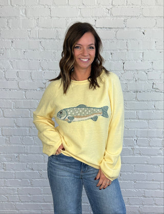 Woman wearing a butter yellow sweater with a blue and brown trout on the front paired with medium washed denim, cuffed jeans, and brown shoes.