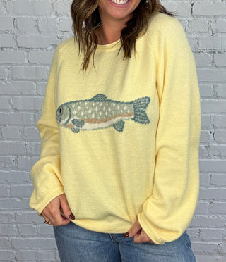 Woman wearing a butter yellow sweater with a blue and brown trout on the front paired with medium washed denim, cuffed jeans, and brown shoes.