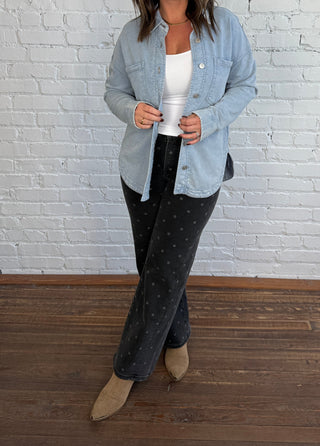 Woman wearing a denim button up shirt with a white undershirt and black jeans with grey polkadots and taupe boots.