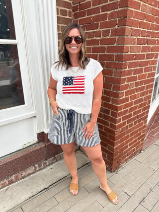 Woman wearing a white t-shirt with an American flag design and striped shorts, standing against a brick wall.