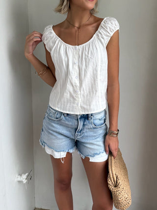 Woman wearing a white top and denim shorts standing against a plain wall.